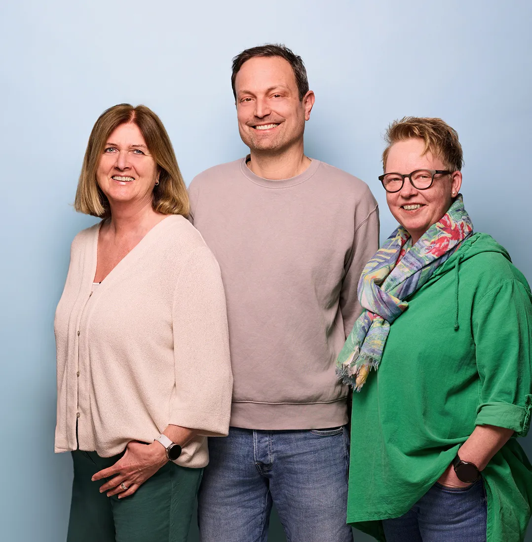 Drei lächelnde Personen stehen nebeneinander vor einem hellblauen Hintergrund.